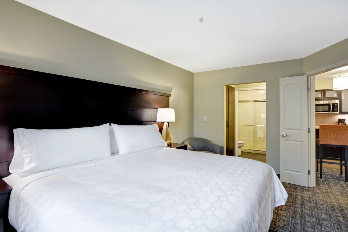 A cozy suite interior featuring a king bed with plush linens and a lamp, a doorway leading to a bathroom, and a kitchenette area—all set within a comfortably arranged, individualized decor. The overall atmosphere is warm and homey, inviting relaxation and convenience.