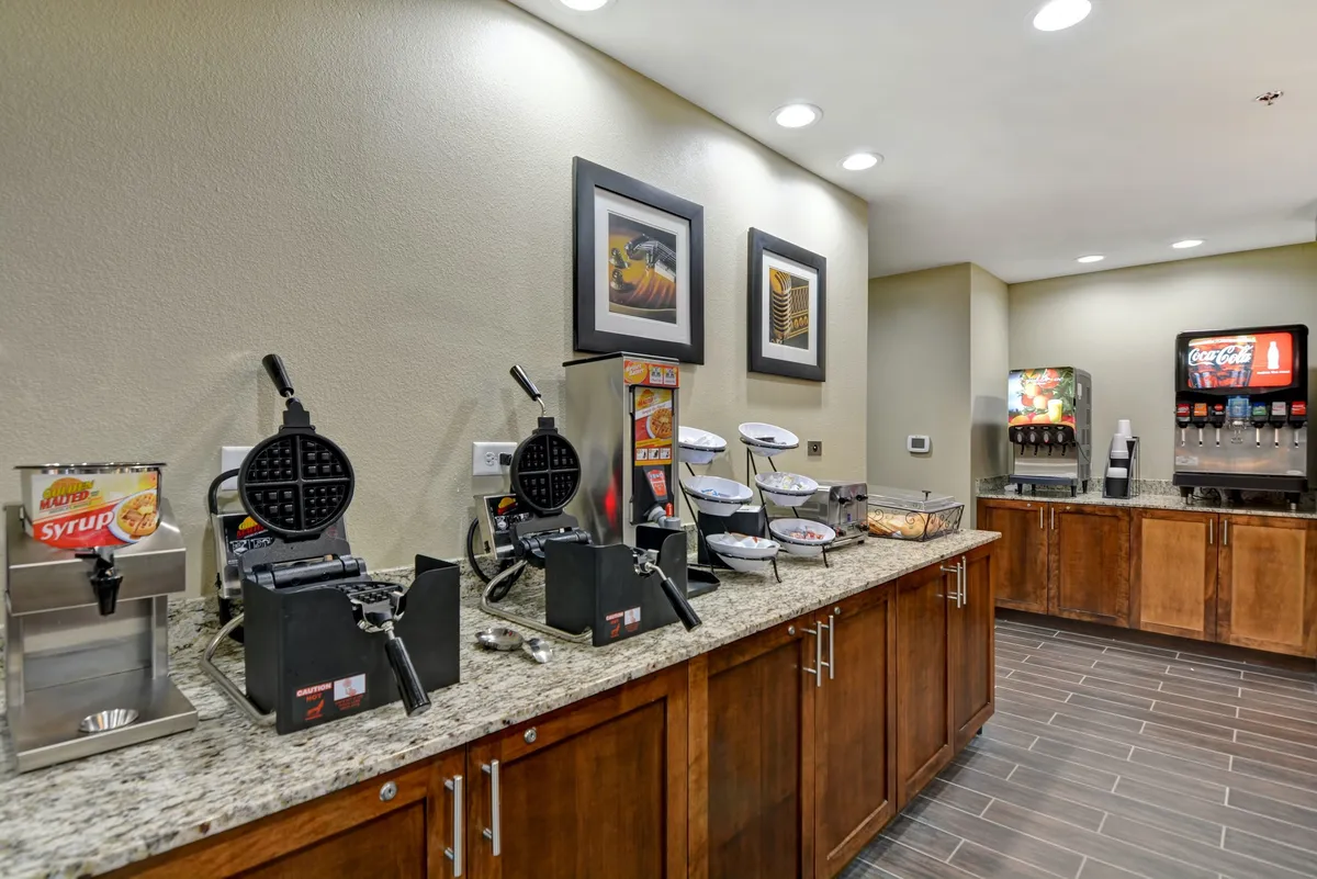 A bright breakfast area featuring waffle makers, a syrup dispenser, a beverage station, and lively wall art. Guests are seen preparing their breakfast and socializing at the beverage counter, giving the space a friendly and approachable feel.