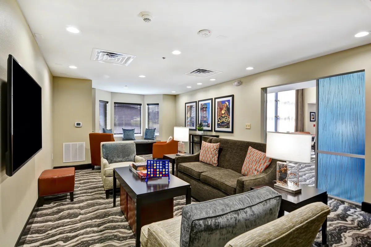 A bright living area furnished with a sofa adorned with patterned cushions, a coffee table set up for board games, a wall-mounted TV, and colorful accent chairs. Decorative artwork adds character, and the space feels lively yet comfortable, perfect for relaxing or socializing.