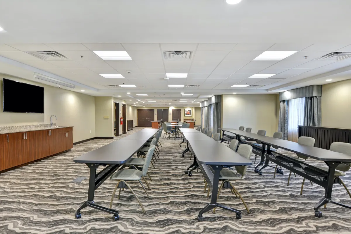 A spacious business center with long tables arranged in a U-shape, a large TV screen for presentations, and ample cabinet storage. The atmosphere is collaborative and practical, suitable for productivity and group meetings.