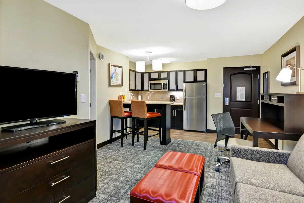 This image showcases a cozy, modern condo interior featuring a compact kitchen with stainless appliances, a pair of inviting bar stools at the counter, a plush ottoman, and a large flat-screen TV—the perfect setup for relaxing nights in or casual meals together.