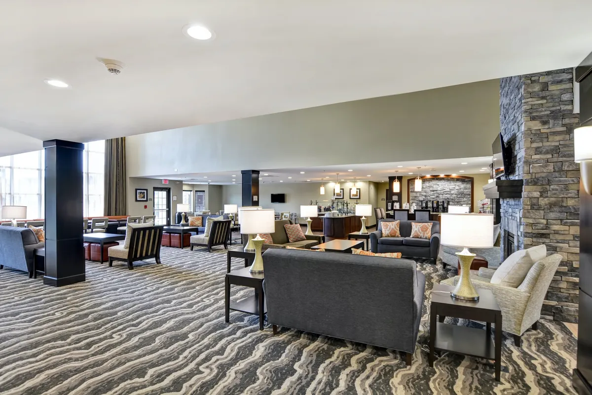 This image features an inviting hotel lobby with a fireplace as its centerpiece, surrounded by comfortable seating, a lobby desk, and large windows that let in ample natural light. Guests can be seen socializing or relaxing, creating a lively yet comfortable atmosphere that serves as a welcoming communal space.
