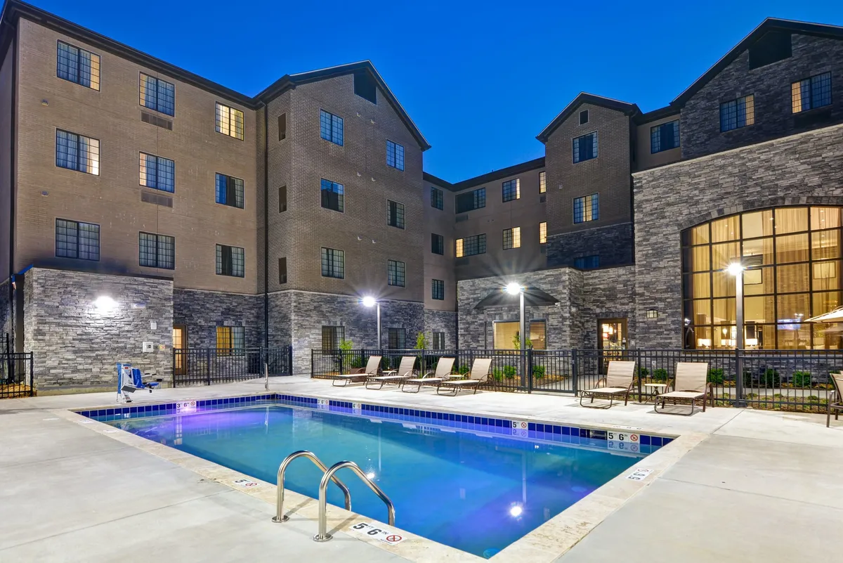 A wide exterior shot reveals the building facade with a sparkling swimming pool in the foreground and comfortable lounge chairs arranged for relaxation. The scene is set during a relaxed, social time, ideal for unwinding after a workday or connecting with colleagues outdoors.