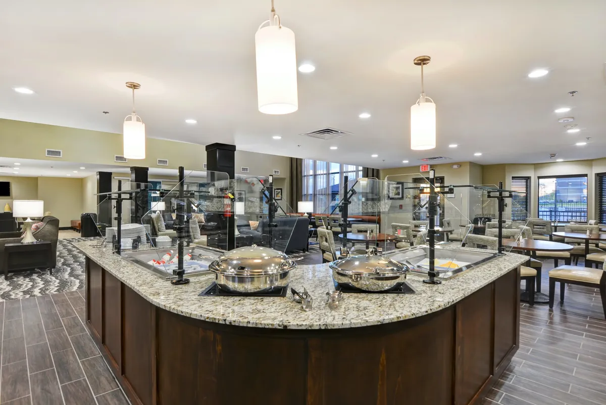 A view of a modern kitchen with a granite countertop, serving dishes, and inviting lighting. The kitchen is connected to a seating area, suggesting a space designed for both cooking and gathering. The setup is clean and ready for meal preparation or relaxed dining.