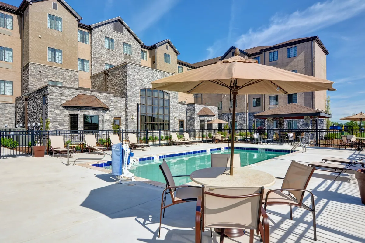 An exterior view of the hotel’s seasonal outdoor pool, surrounded by sun loungers and sun umbrellas. Guests are relaxing by the pool and enjoying the sunshine, while the building's facade provides a welcoming backdrop.