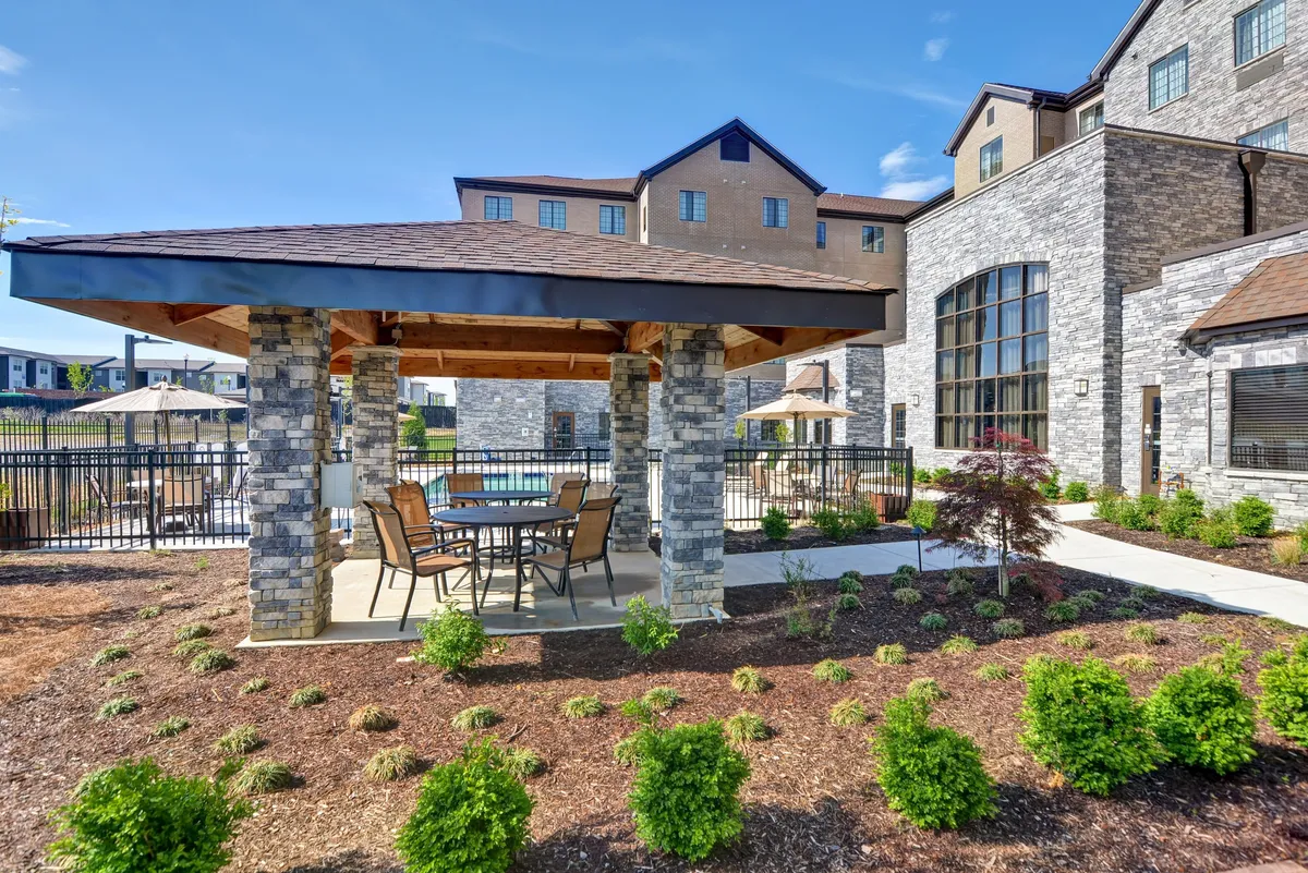 A beautifully landscaped outdoor area features a stone gazebo with comfortable seating, surrounded by greenery and a swimming pool visible in the background. The tranquil atmosphere encourages relaxation, casual meetings, or quiet moments between work obligations.
