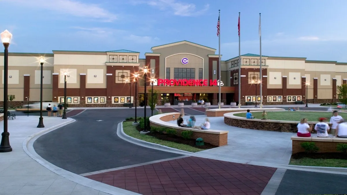 The illuminated facade of a cinema at Providence Marketplace is shown at dusk, with vibrant lights, surrounding plaza, and nearby flag poles. The scene pulses with life, inviting guests to explore local entertainment and community events after work hours.